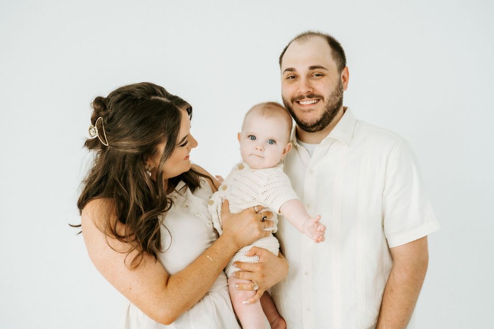 A happy young family with a smiling man and woman holding an adorable baby in white outfits against a light background.