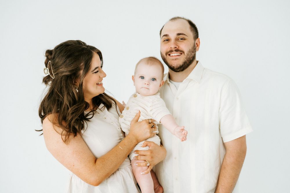 A smiling couple in white outfits holds their baby together against a clean white background.
