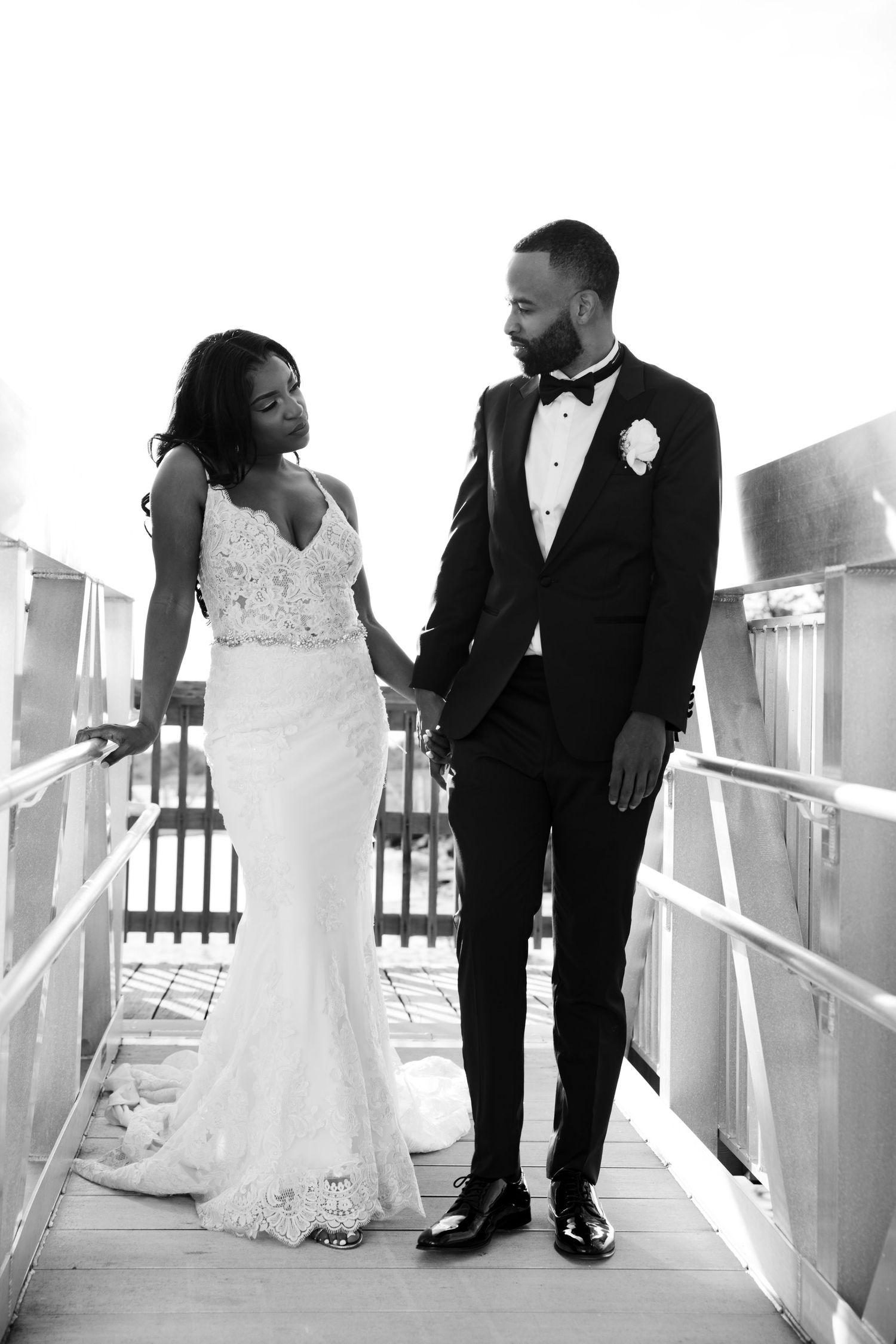 Black and white wedding photo of a bride in lace gown and groom in tuxedo holding hands on a bridge.