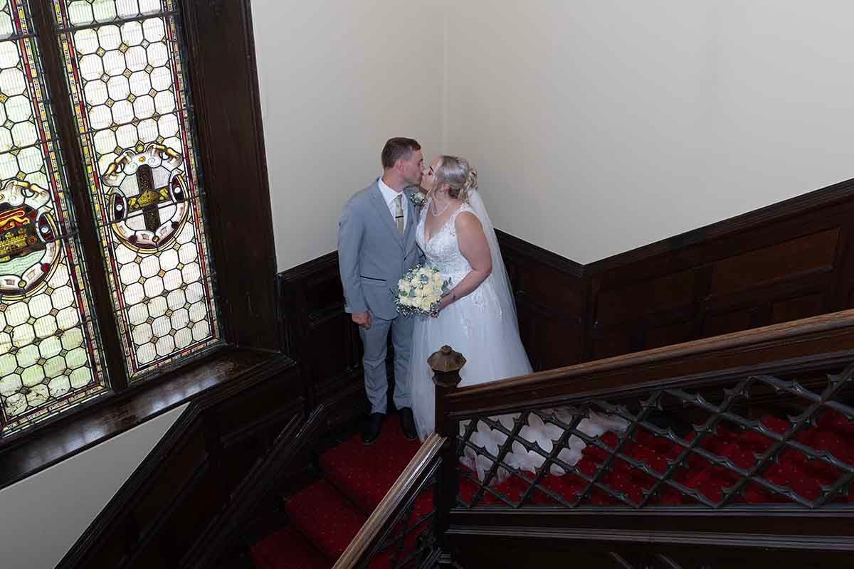 Bride and groom share a kiss on a grand staircase with red carpet and stained glass windows.