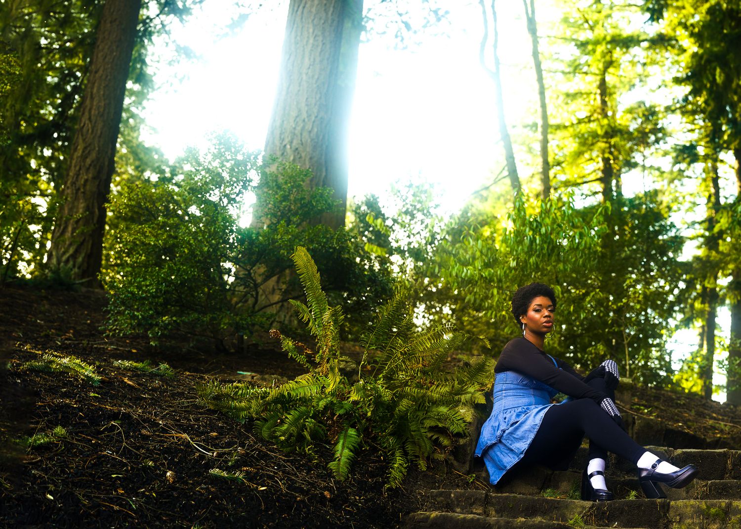 Young woman sitting on mossy stone steps in a lush green forest with sunlight filtering through tall trees.