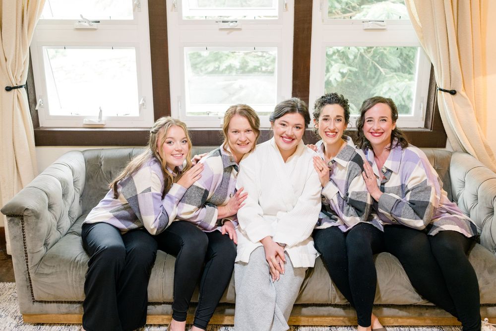 Bride in white robe smiling with four bridesmaids in plaid shawls seated on a gray sofa near a bright window.