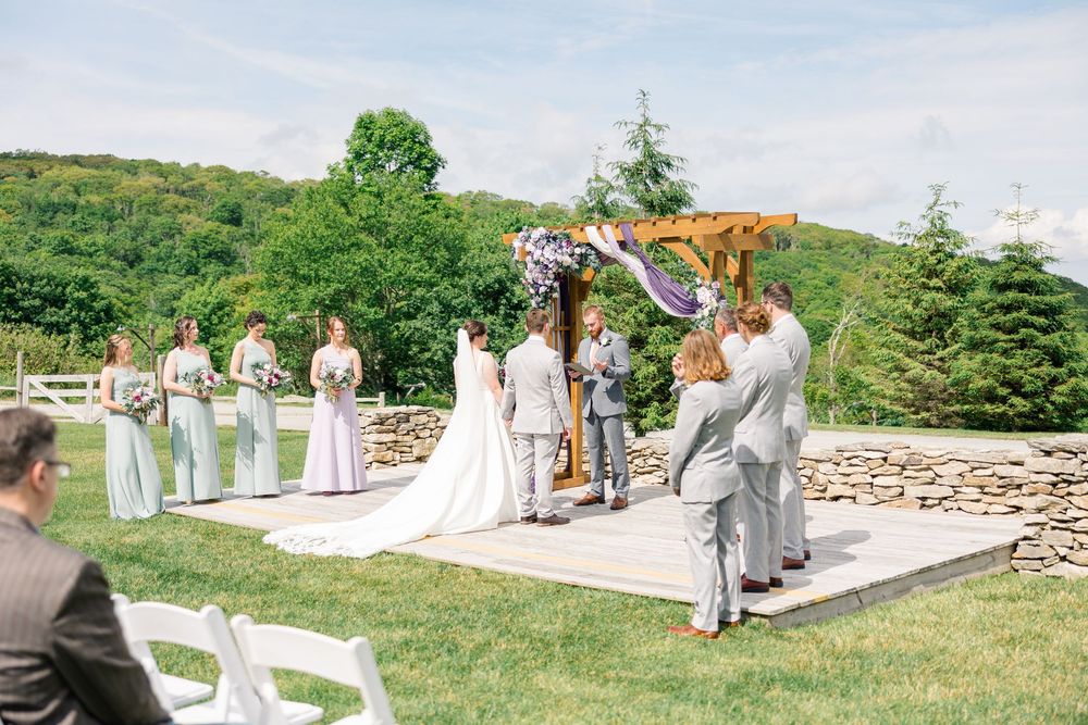 Outdoor wedding ceremony on a wooden platform with bridesmaids, groomsmen, and couple at a floral arch amid lush green hills.