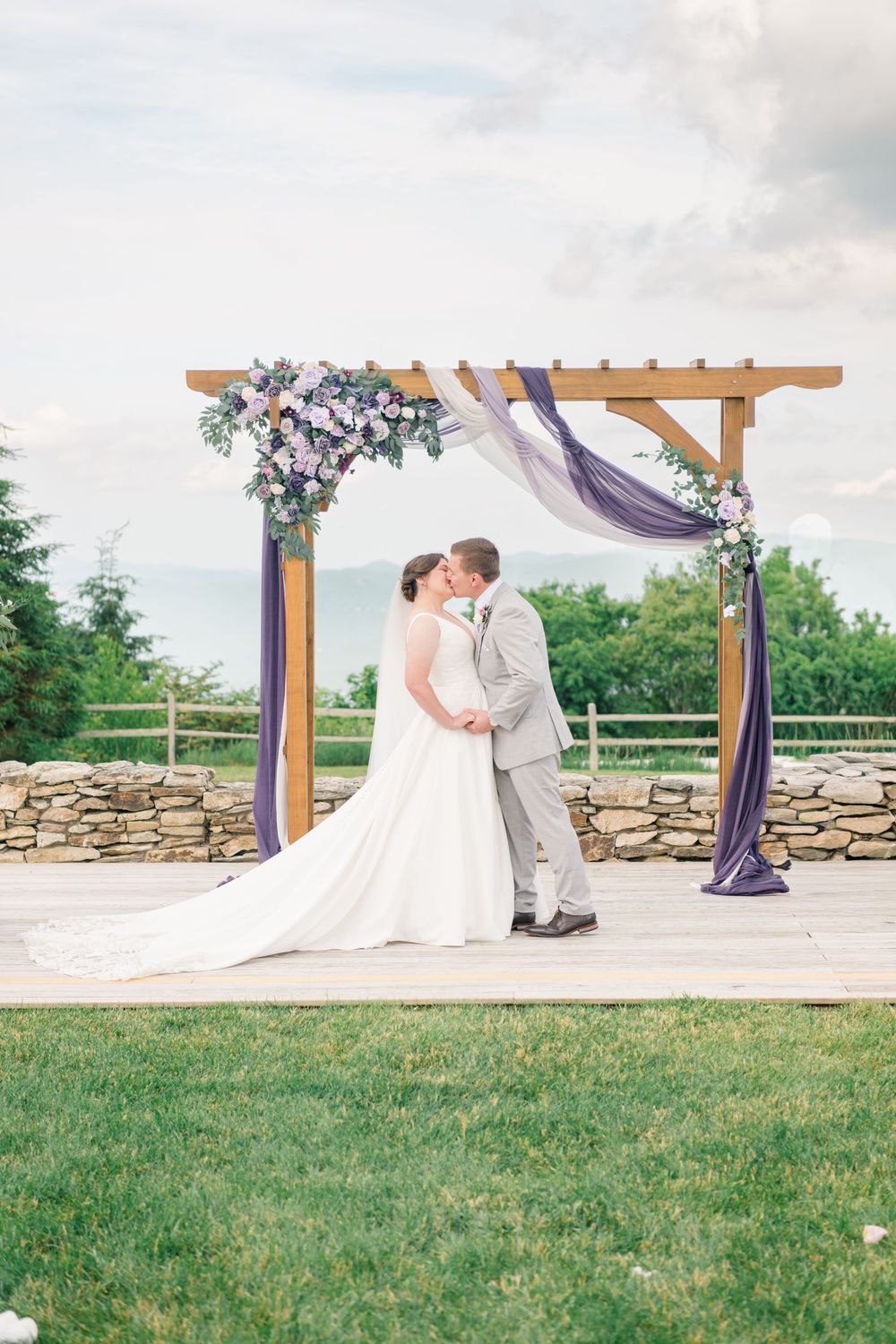 Bride and groom share first kiss under wooden arch with purple draping and floral arrangements at outdoor wedding ceremony.