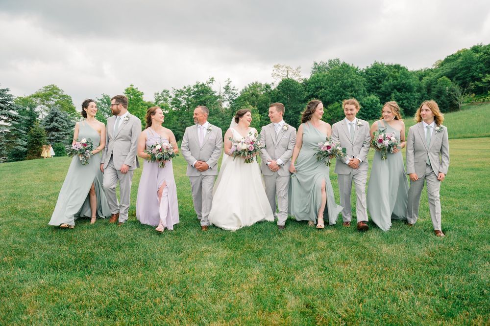 Wedding party walks outdoors on green lawn, bride in white gown with bridesmaids in sage green dresses and groomsmen in gray suits.