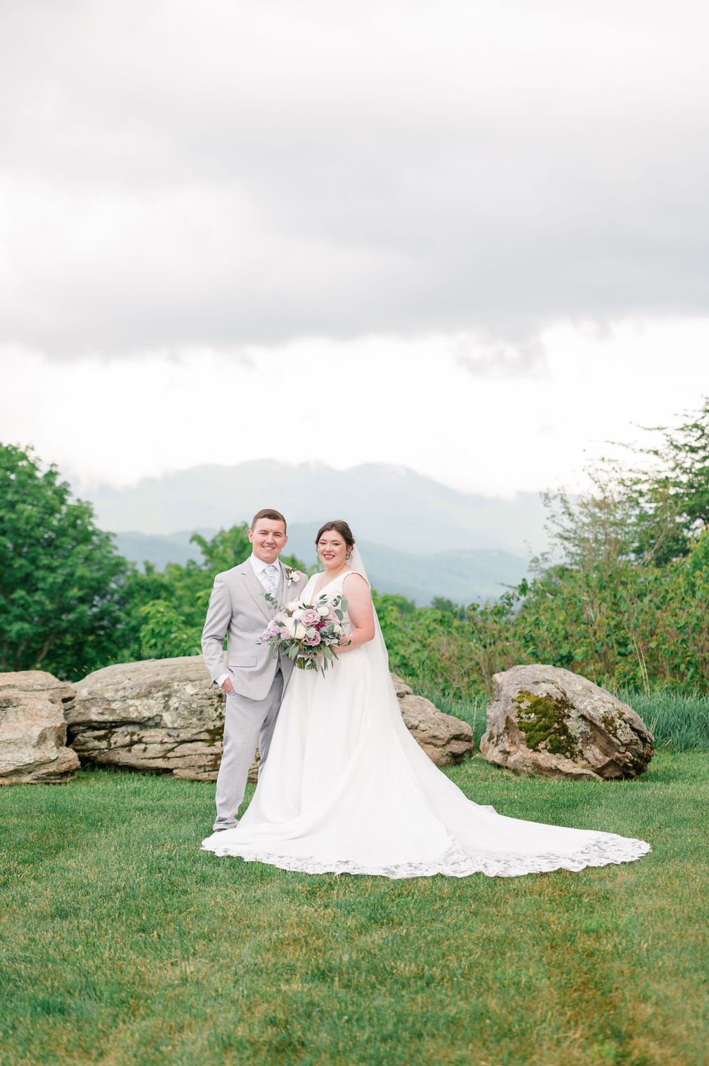Bride and groom pose on green lawn with mountain backdrop, she holds pink bouquet in white gown, he wears gray suit.