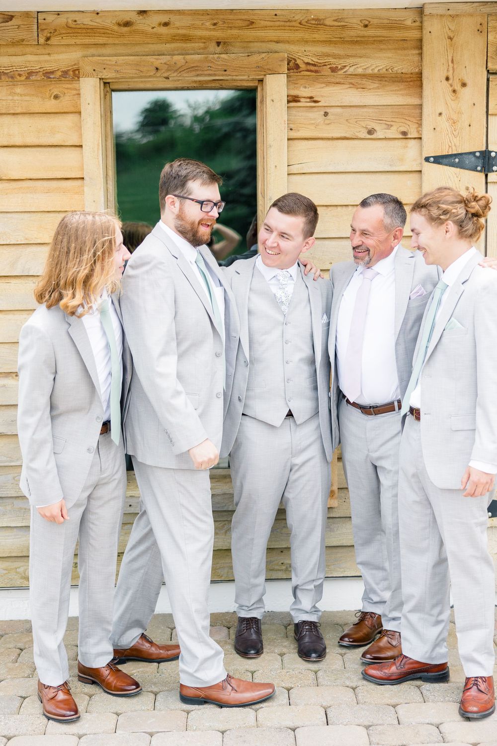 Groom and groomsmen in light gray suits with brown dress shoes laughing together outside a rustic wooden barn.