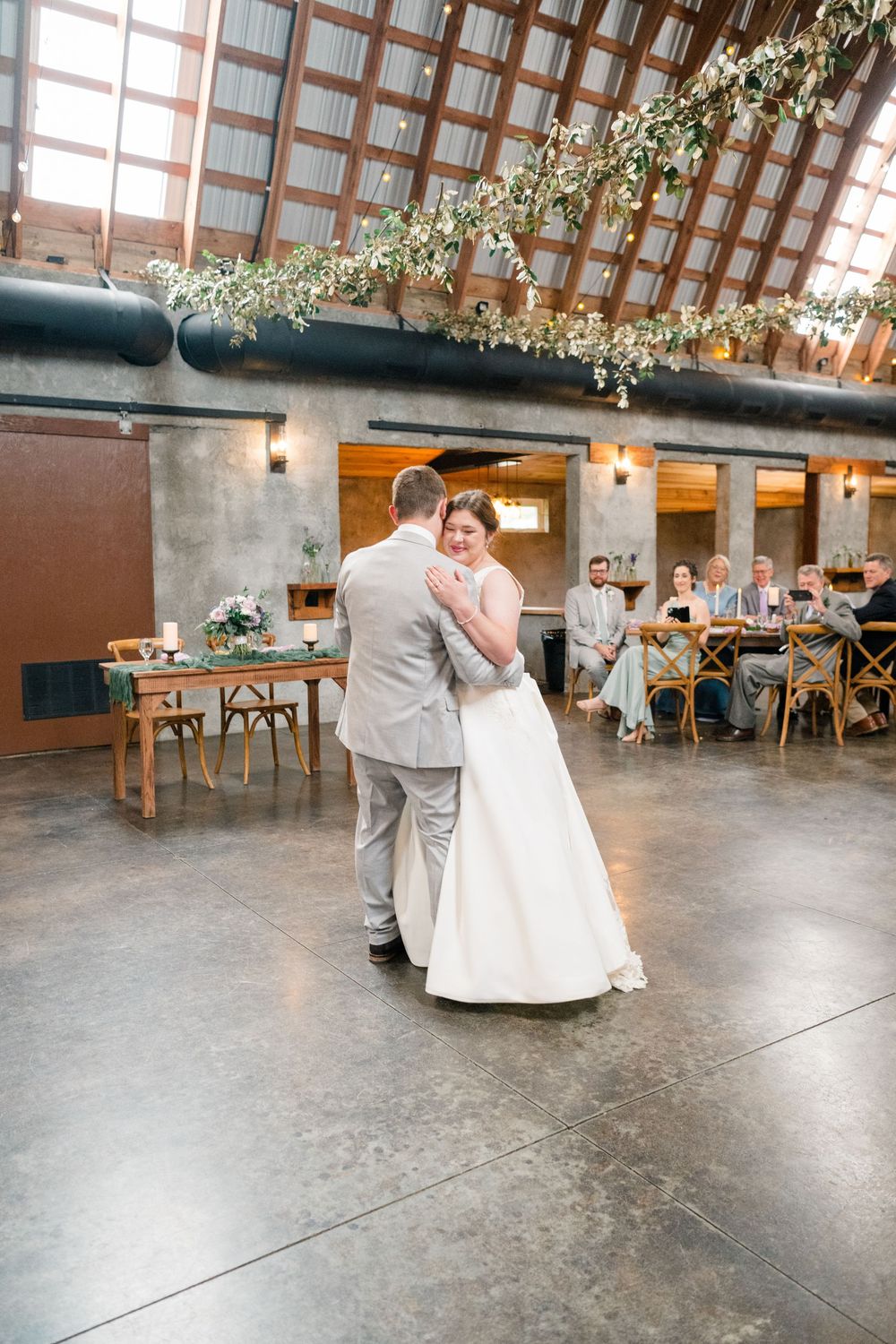 Bride and groom share their first dance in a rustic industrial wedding venue with floral ceiling decor and seated guests.