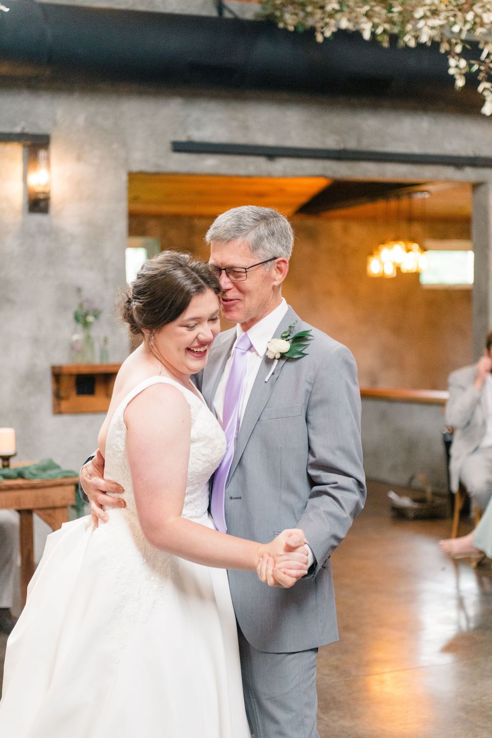 Bride and father share a joyful first dance at wedding reception, both smiling, groom in gray suit with lavender tie.