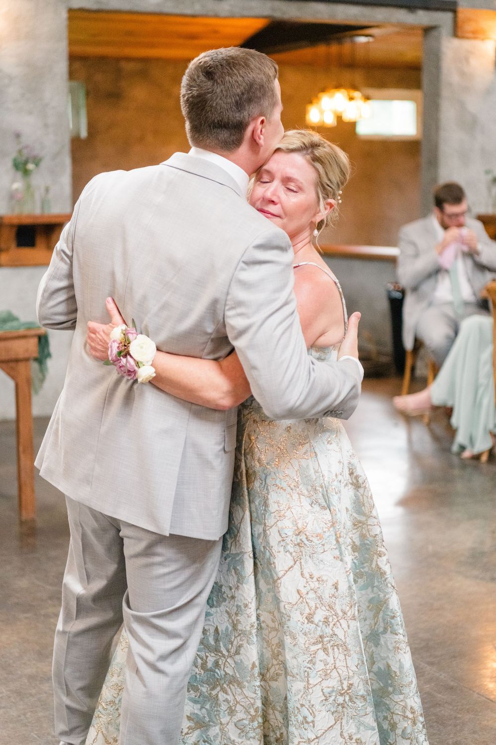 Emotional mother-son wedding dance, woman in floral gown with corsage embraces groom in tan suit.