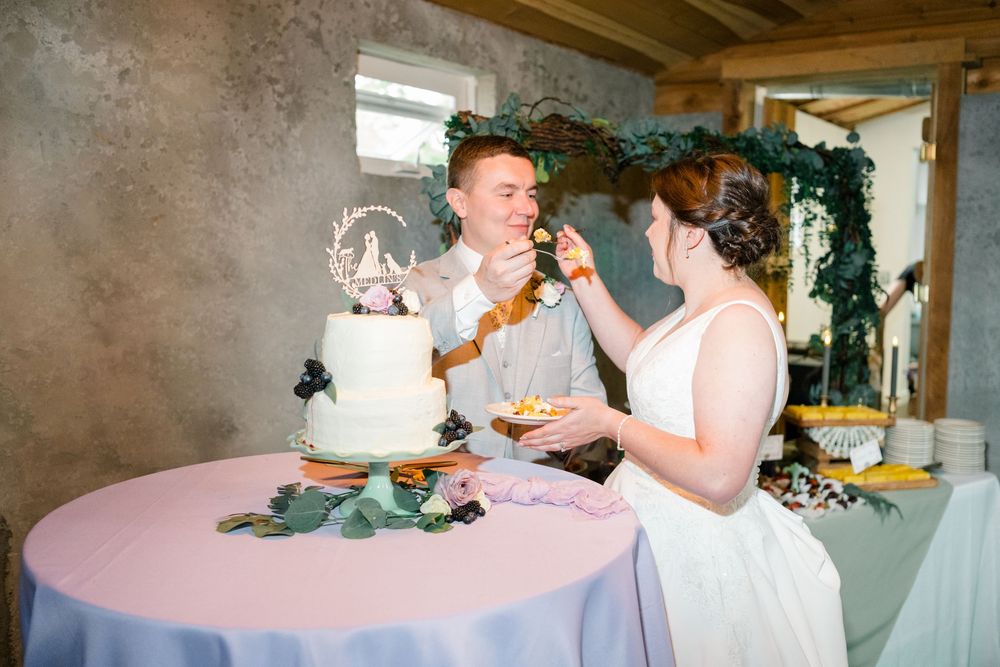 Bride and groom feeding each other wedding cake at reception table with white tiered cake and floral decor.
