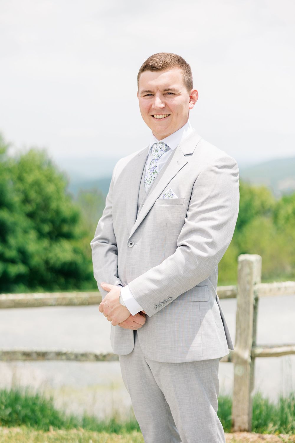 Smiling groom in light gray suit with floral tie stands outdoors near wooden fence with green hills backdrop.