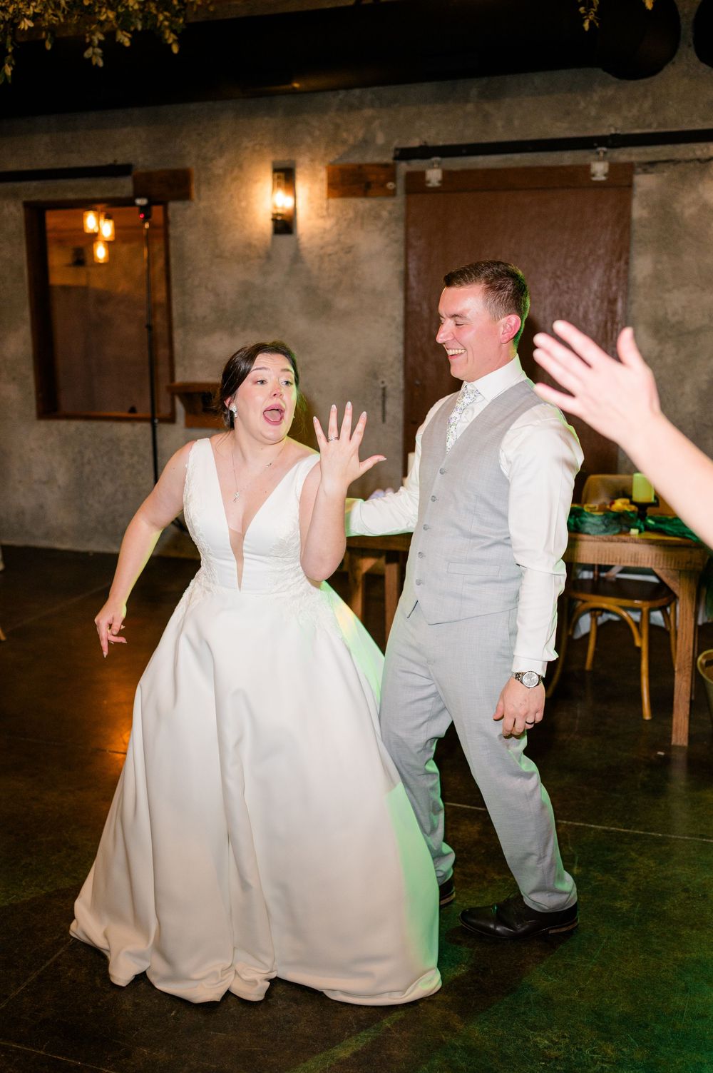 Bride and groom dancing joyfully at their wedding reception in a rustic venue, bride in white gown, groom in gray suit.
