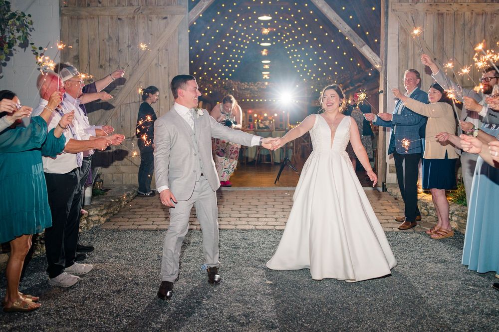 Bride and groom exit wedding reception through sparkler tunnel at rustic barn venue with guests celebrating.