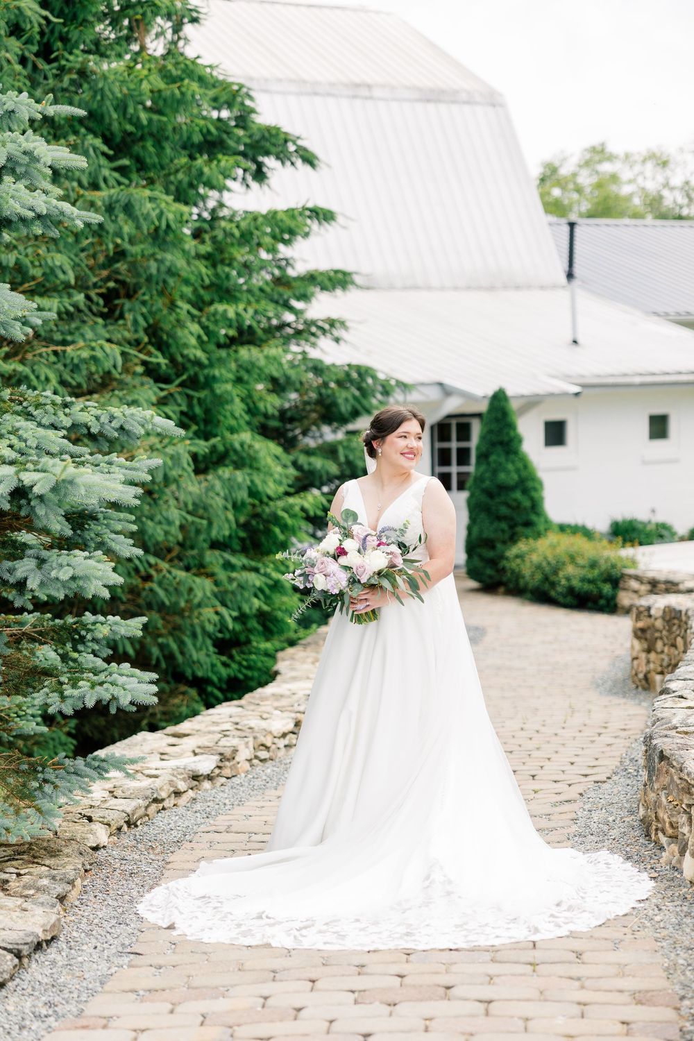 Bride in white gown holding floral bouquet stands on brick path beside evergreen trees near white barn venue.