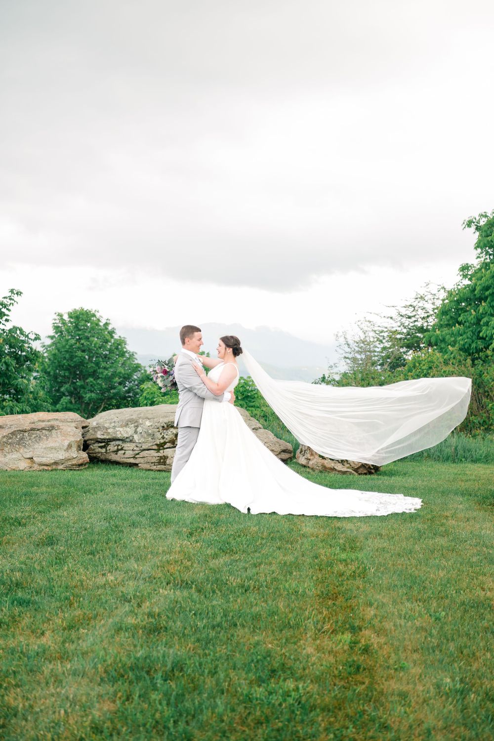 Bride and groom embrace on green lawn with veil flowing in wind, rocks and trees in background on cloudy day.