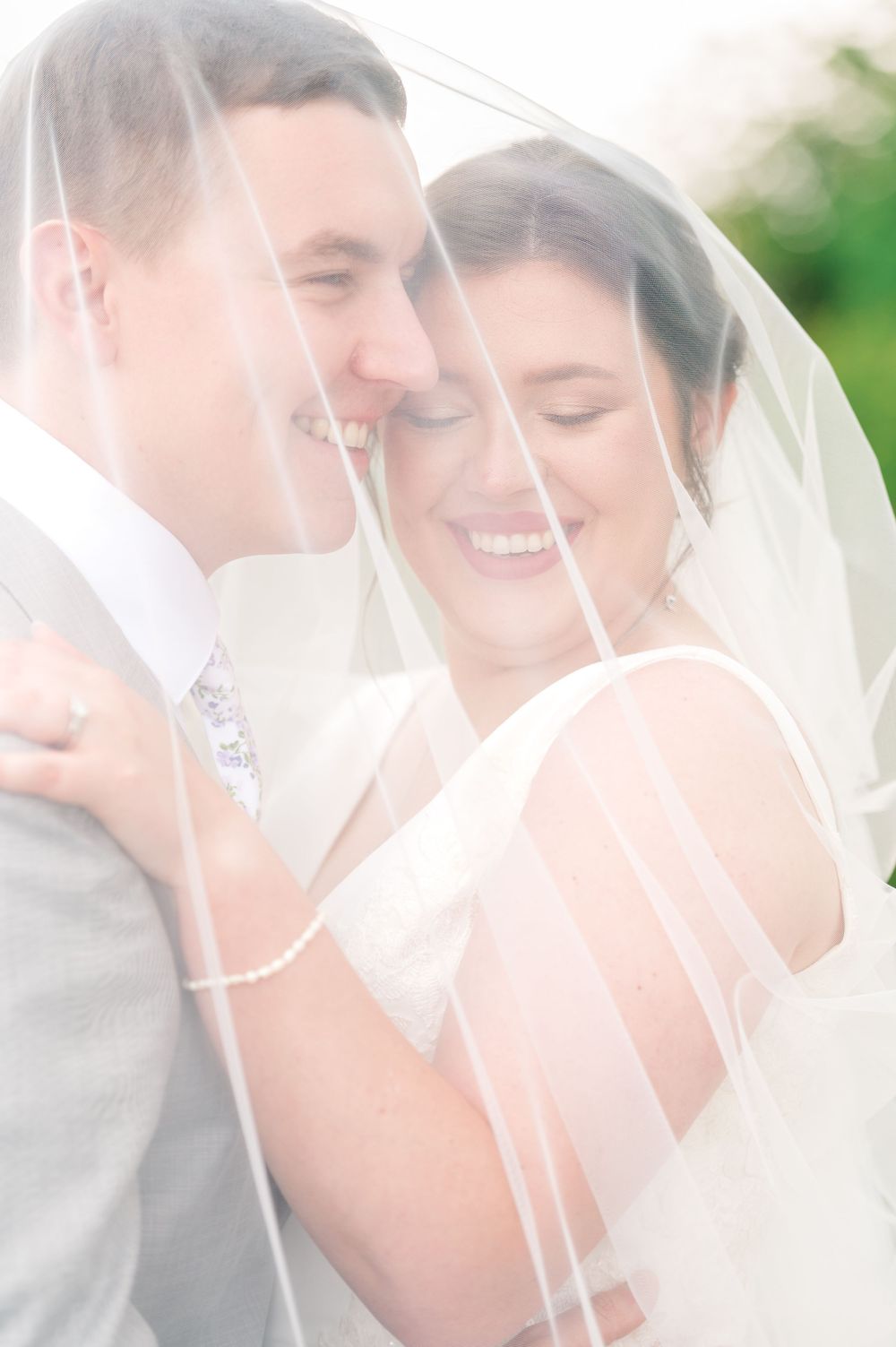Bride and groom smiling together under a sheer wedding veil, sharing an intimate moment on their wedding day.