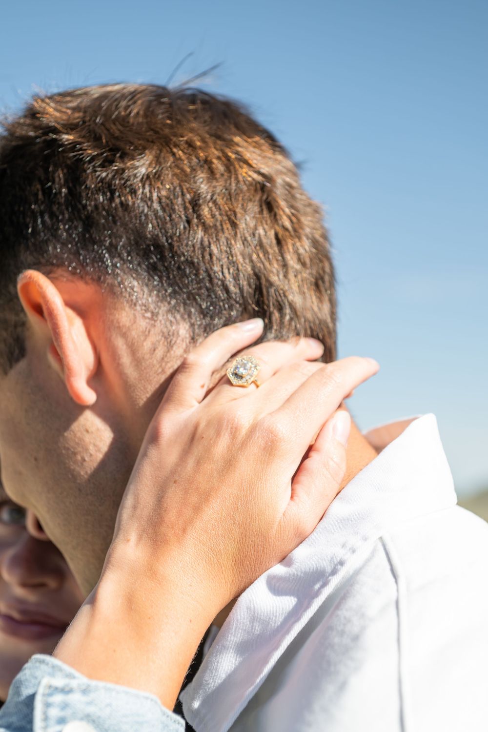 A woman's hand with a gold ring gently rests on the back of a man's neck outdoors under a clear blue sky.