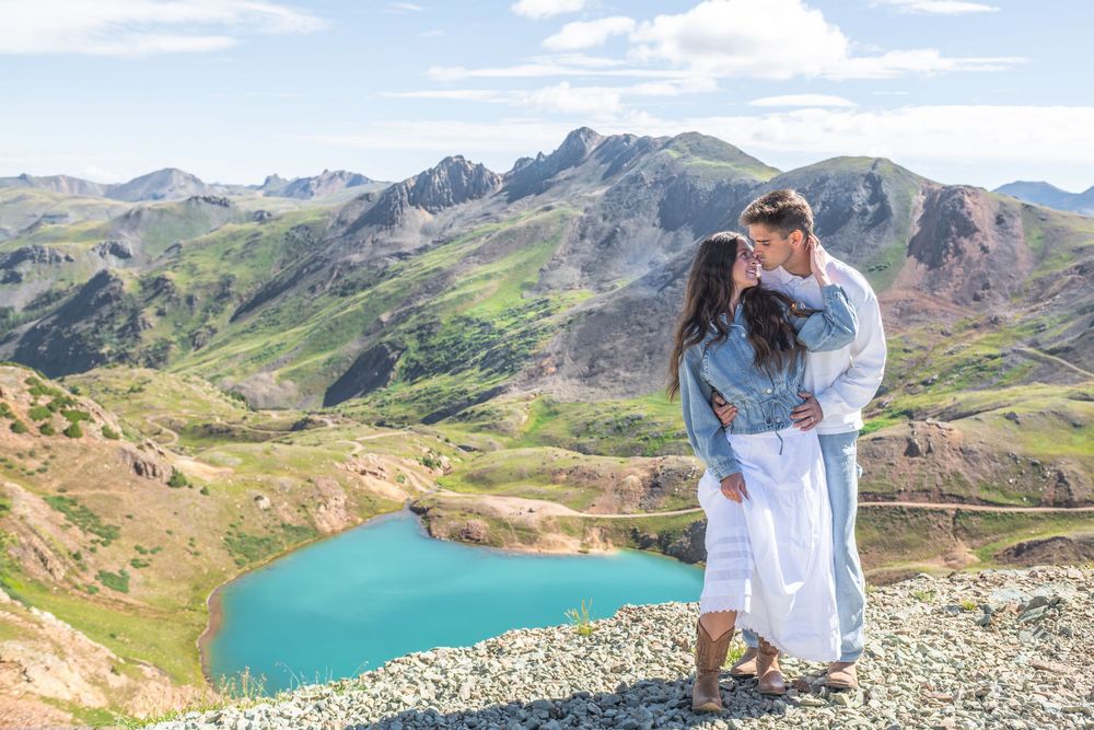 Couple embracing on a rocky mountain overlook with a turquoise alpine lake and green peaks behind.