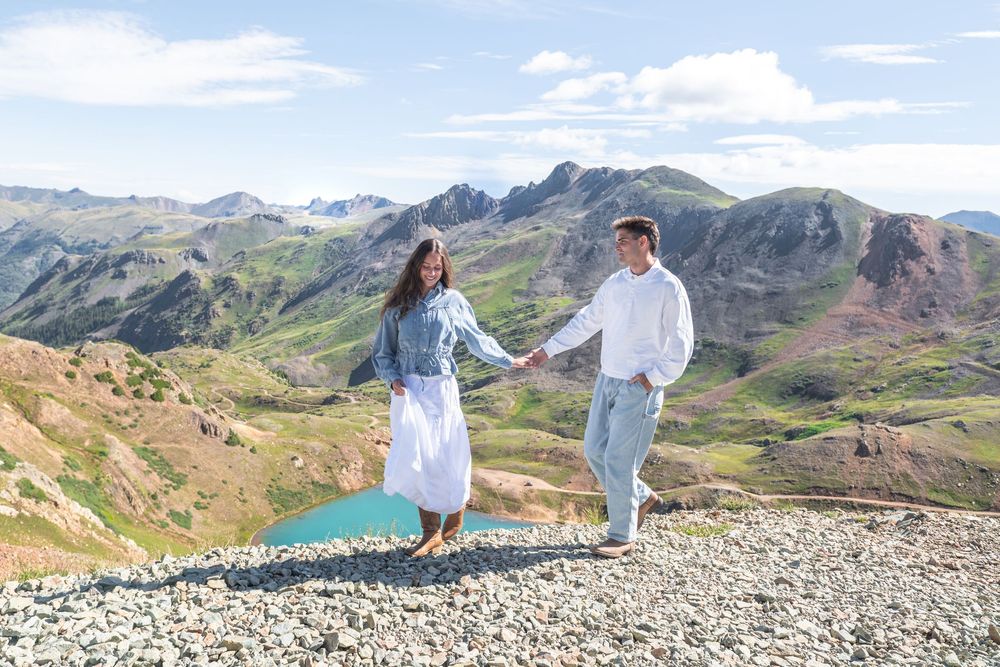 Couple holding hands on mountain summit with scenic green alpine landscape and turquoise lake in background.