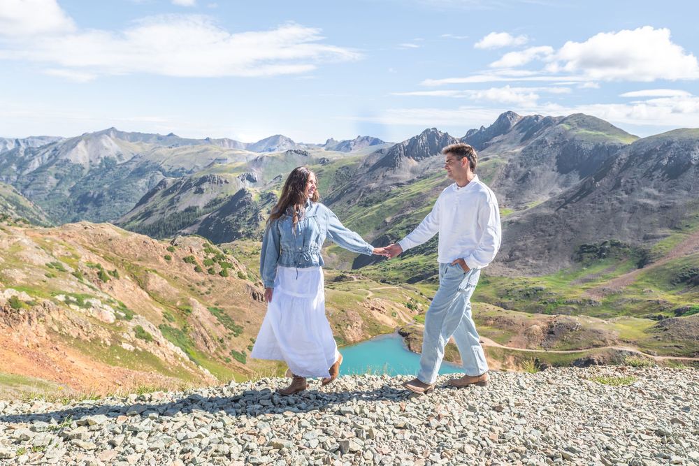 Couple holding hands walking on rocky mountain terrain with turquoise lake and scenic peaks in background.