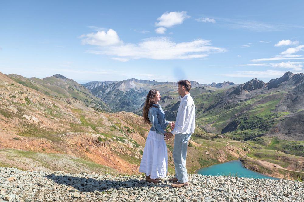 Couple holding hands facing each other with turquoise alpine lake and green mountains backdrop on sunny day.