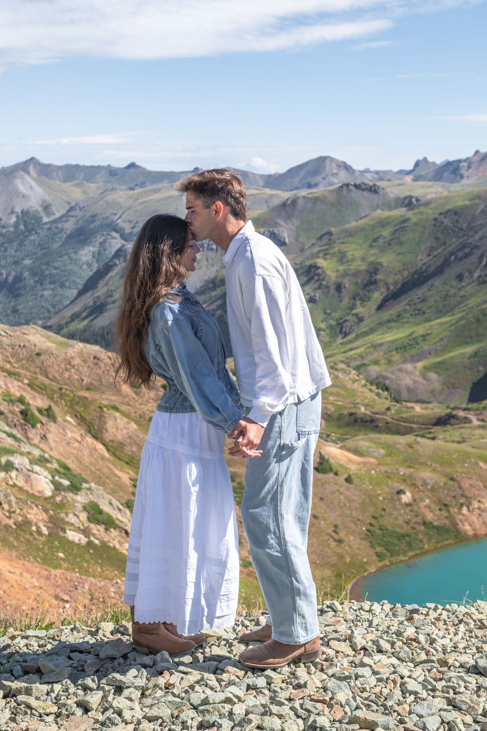 Couple sharing a romantic kiss while holding hands on a rocky mountain overlook with a turquoise lake below.