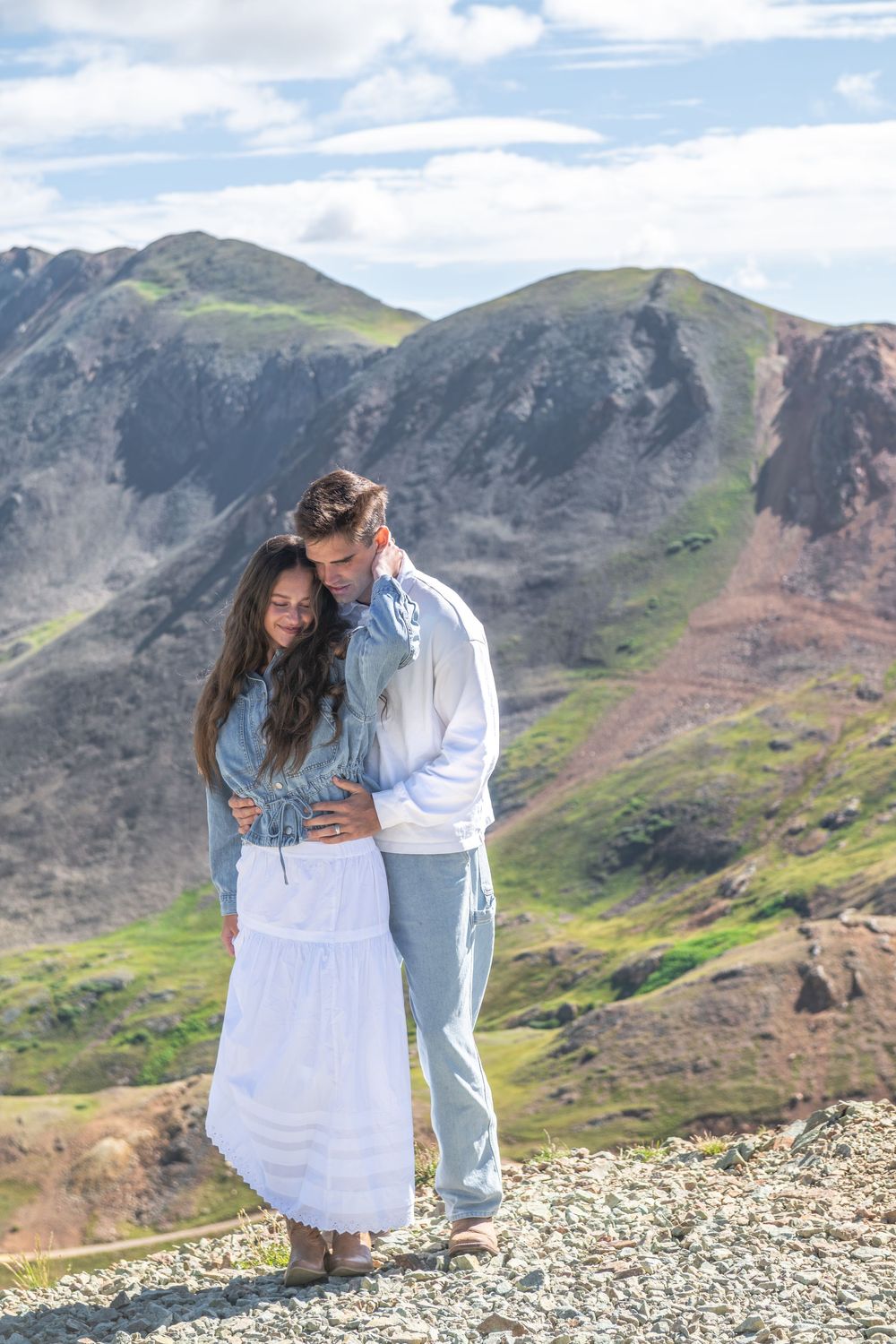 Couple embracing on rocky mountain summit, woman in white skirt and blue top, man in white shirt, green mountains behind.