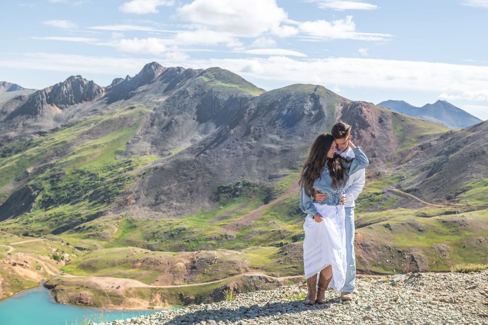 Couple embracing and kissing on a rocky mountain summit with lush green peaks and a turquoise lake below.