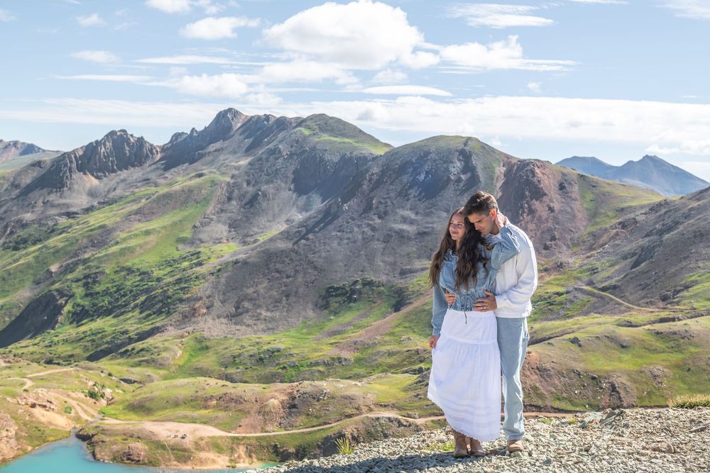 Couple embracing on rocky mountain summit with stunning green alpine valleys and blue lake below on a sunny day.
