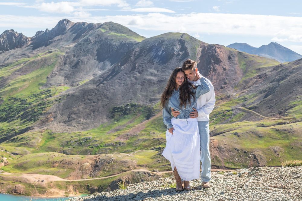 Couple embracing on rocky mountain overlook with green hills and alpine lake in background.