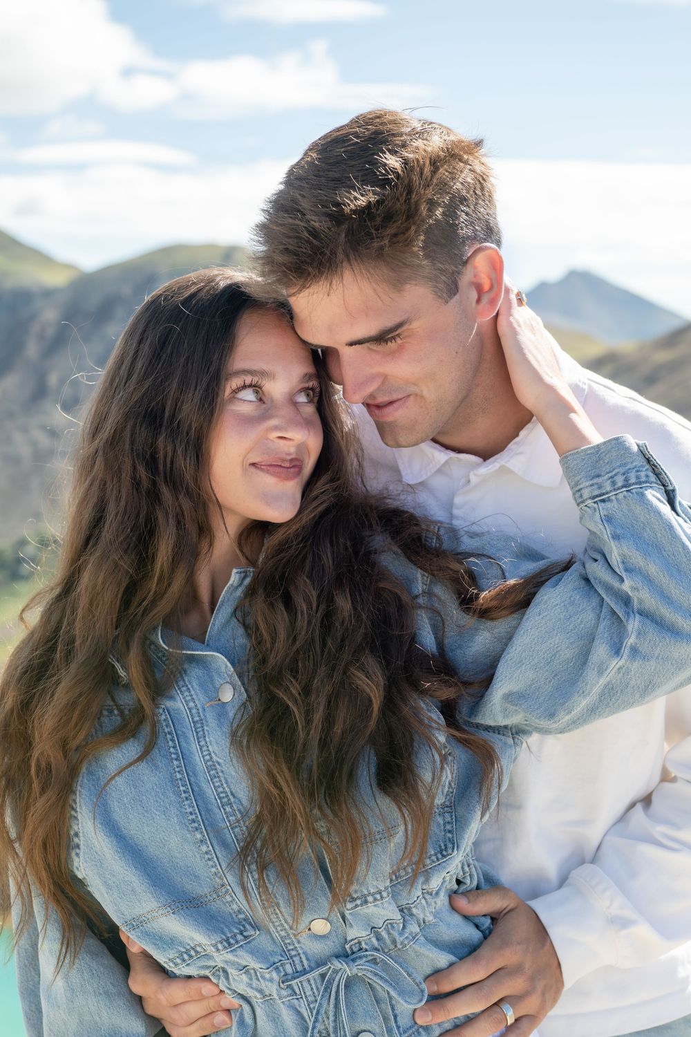 Couple embracing outdoors with mountain backdrop, woman in denim jacket smiling at man in white shirt.