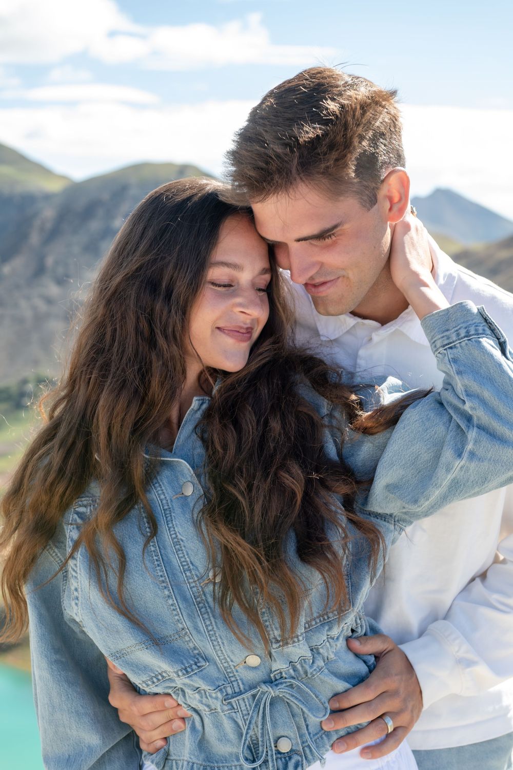 Couple embracing tenderly with mountain lake backdrop, woman in denim jacket, man in white shirt.