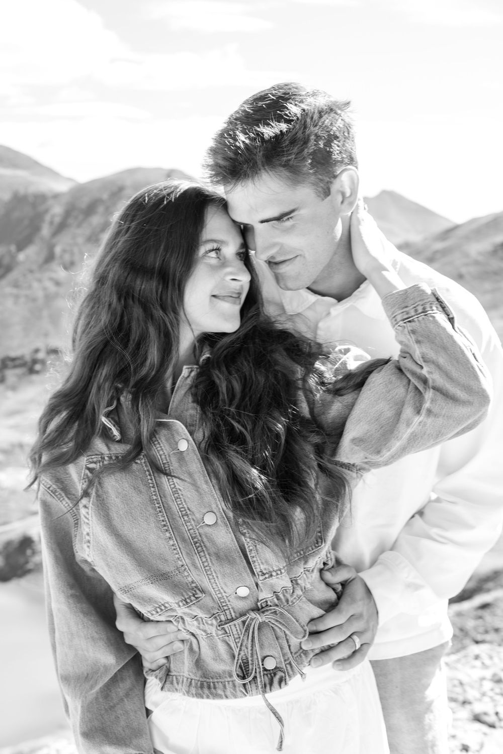 Black and white photo of a romantic couple embracing outdoors with mountains in the background.