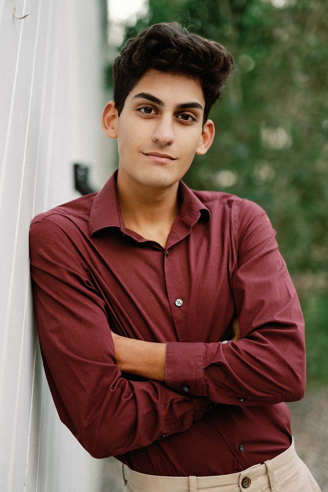 Young person in a burgundy button-down shirt and khaki pants poses confidently against a white wall with greenery in background.