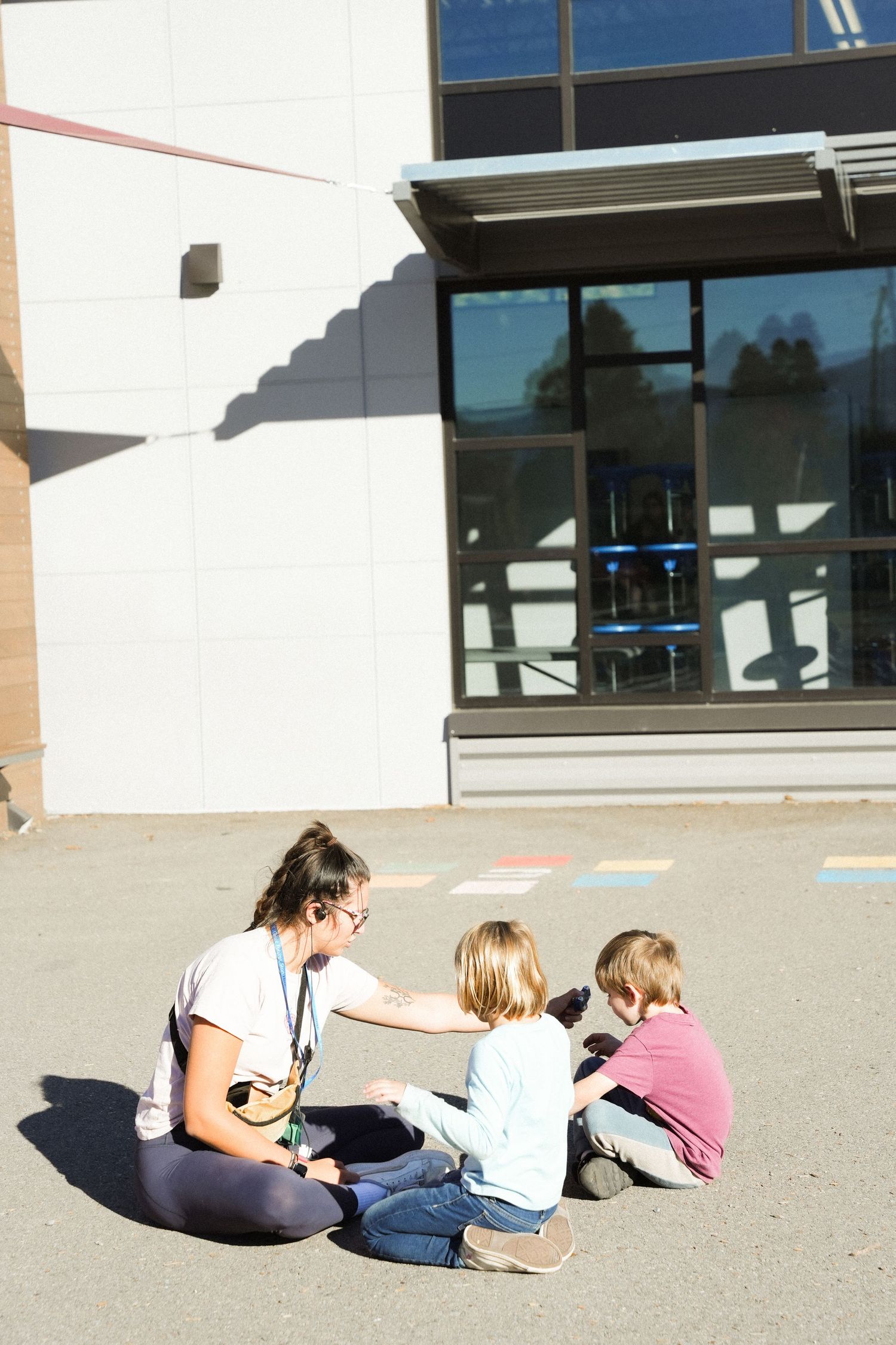 Teacher sitting on pavement with two young students outside a modern school building on a sunny day.
