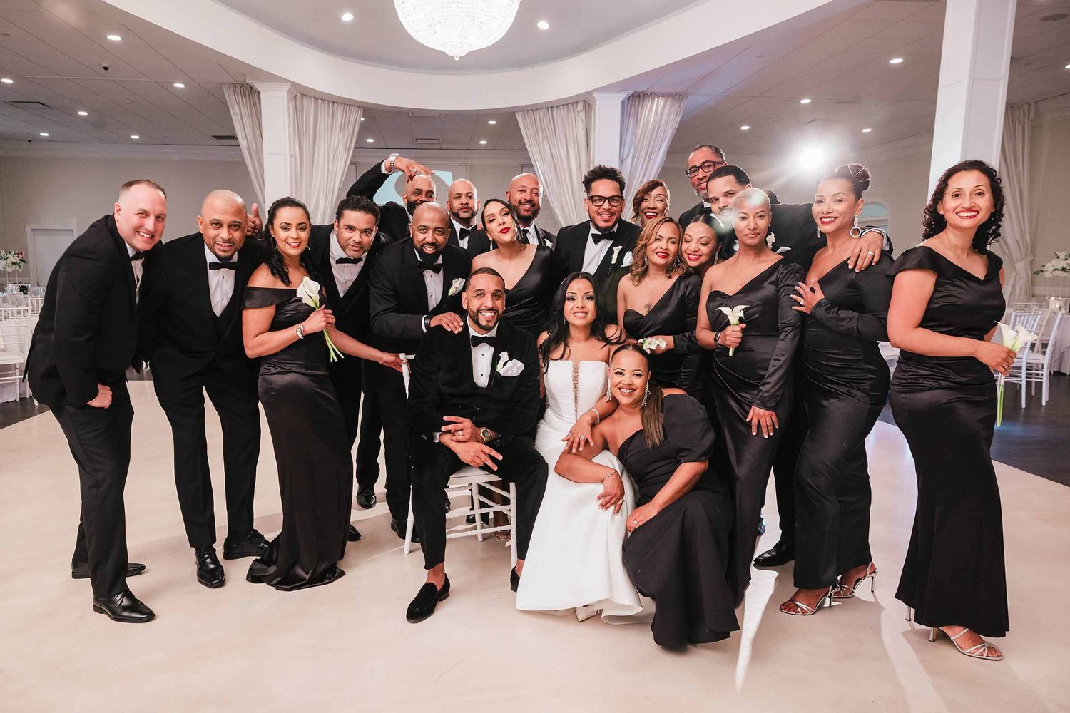 Bride and groom seated with a large formal wedding party in black tie, gathered together in a white ballroom.