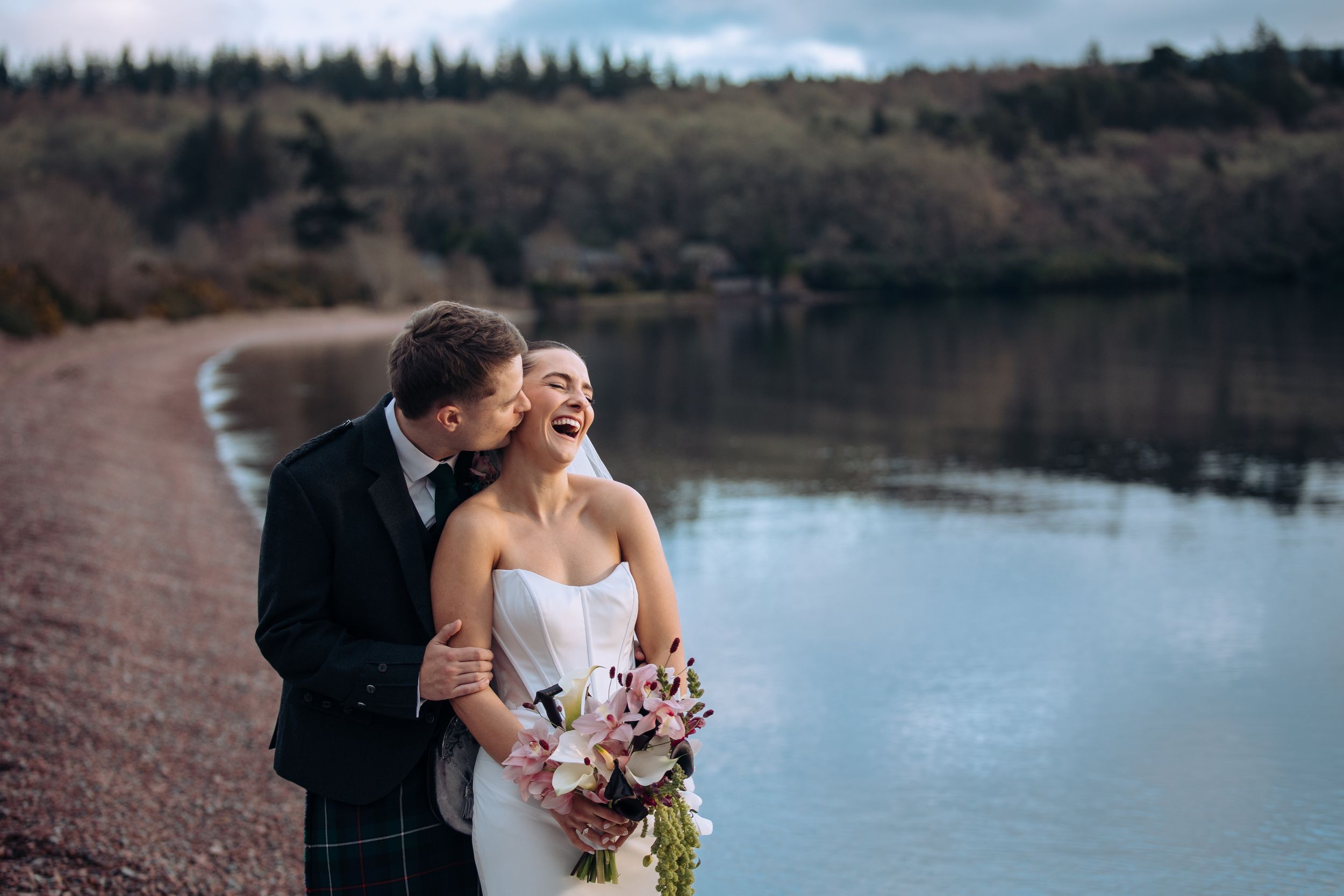 Bride and groom laughing together by a scenic loch in Scotland, groom wearing tartan kilt, bride holding pink bouquet.