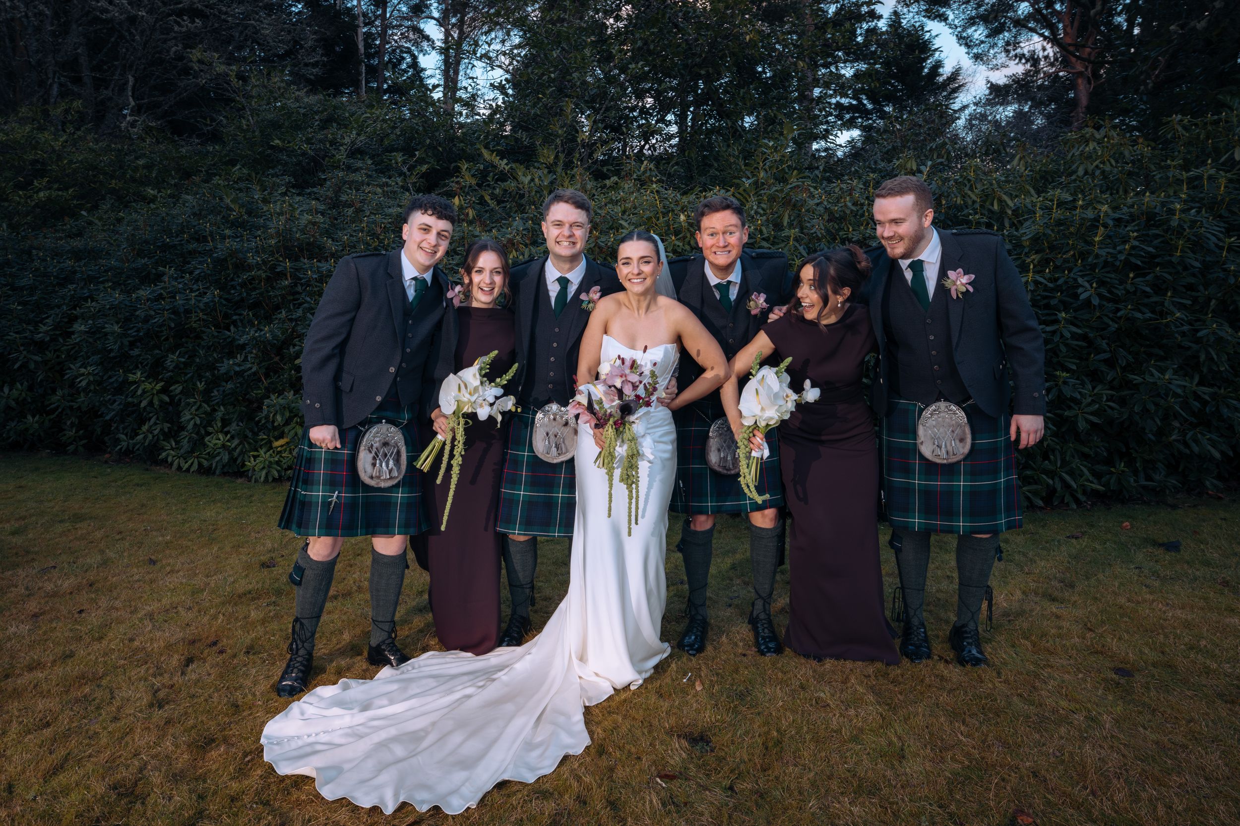 Wedding party posing outdoors, bride in white gown with groomsmen in kilts and bridesmaids in burgundy dresses.