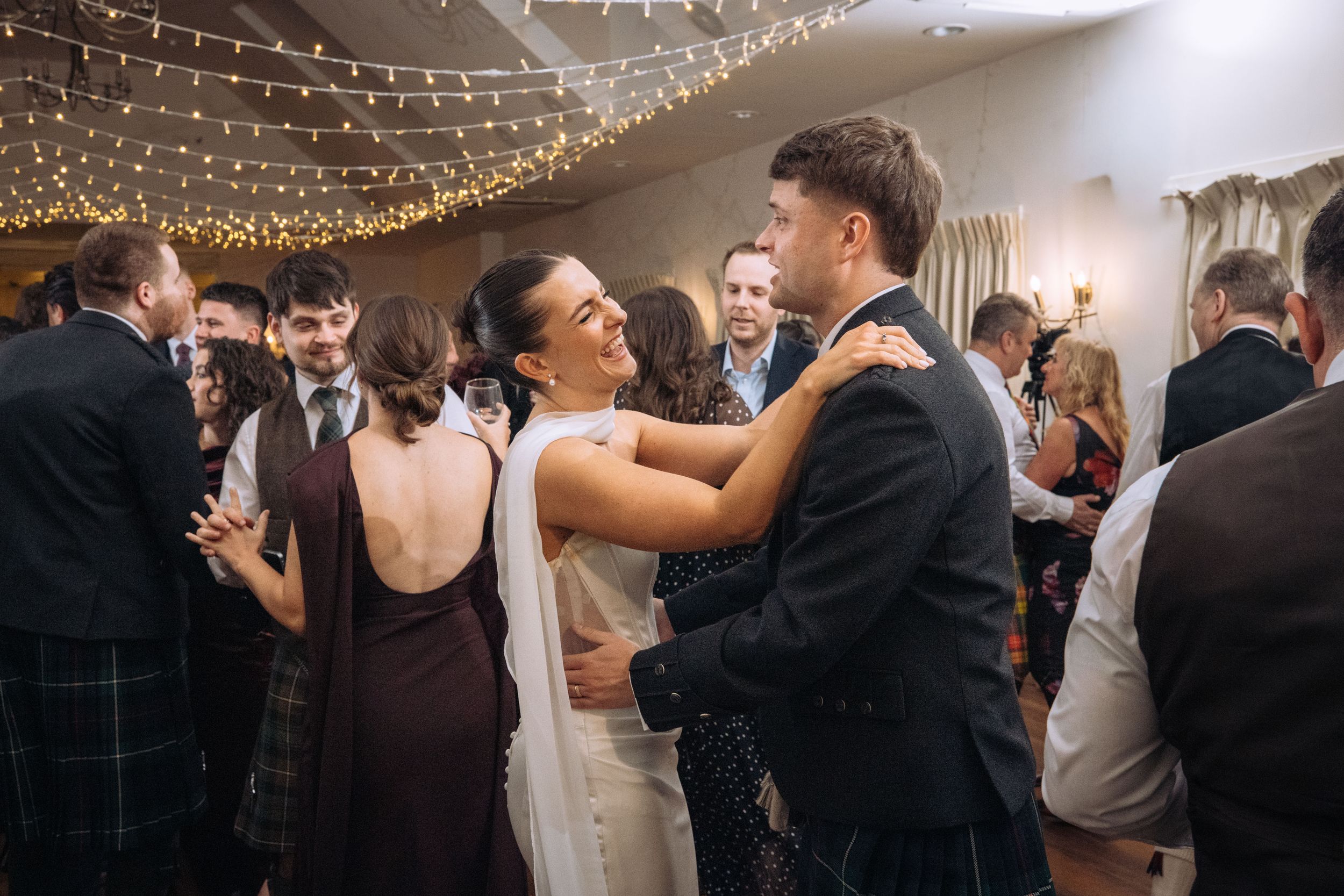 Couple shares a joyful dance embrace at a wedding reception with fairy lights and guests in background.
