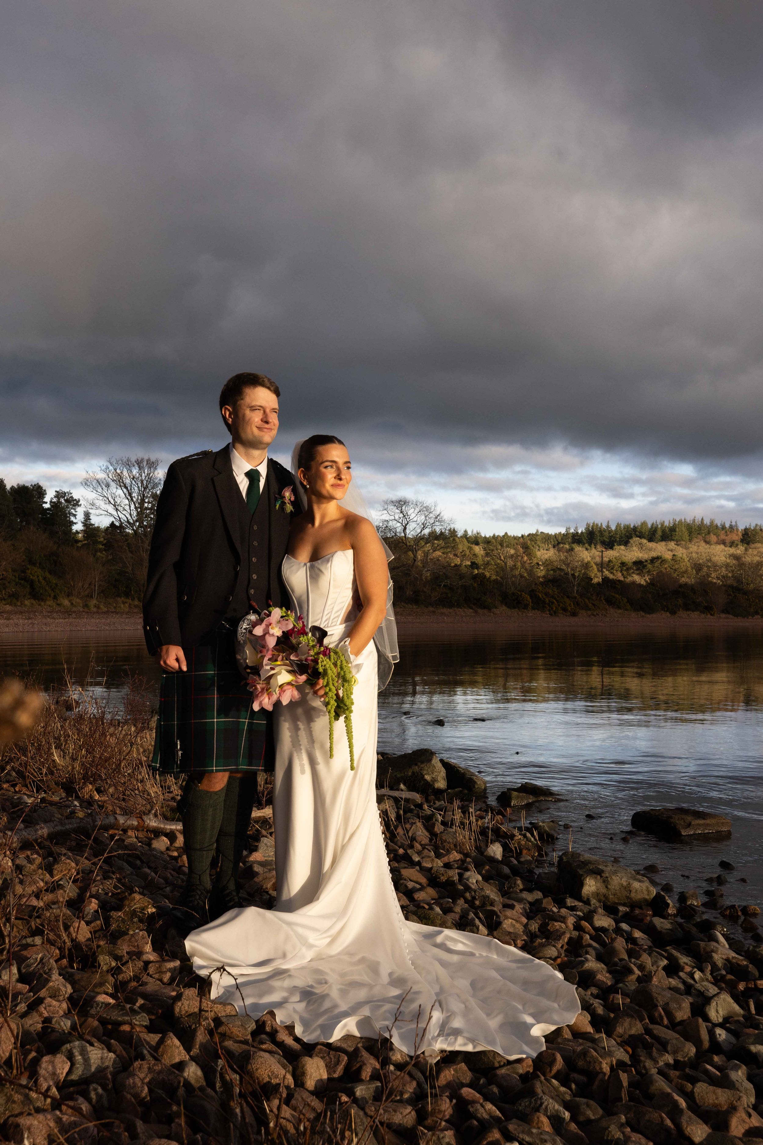 Bride in white gown and groom in Scottish kilt stand on rocky shore by lake under dramatic stormy sky.