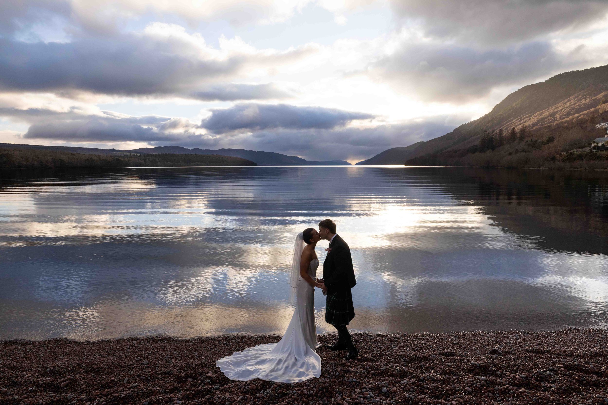 Bride and groom kissing by a serene Scottish loch at sunset, with dramatic clouds reflecting on calm water.