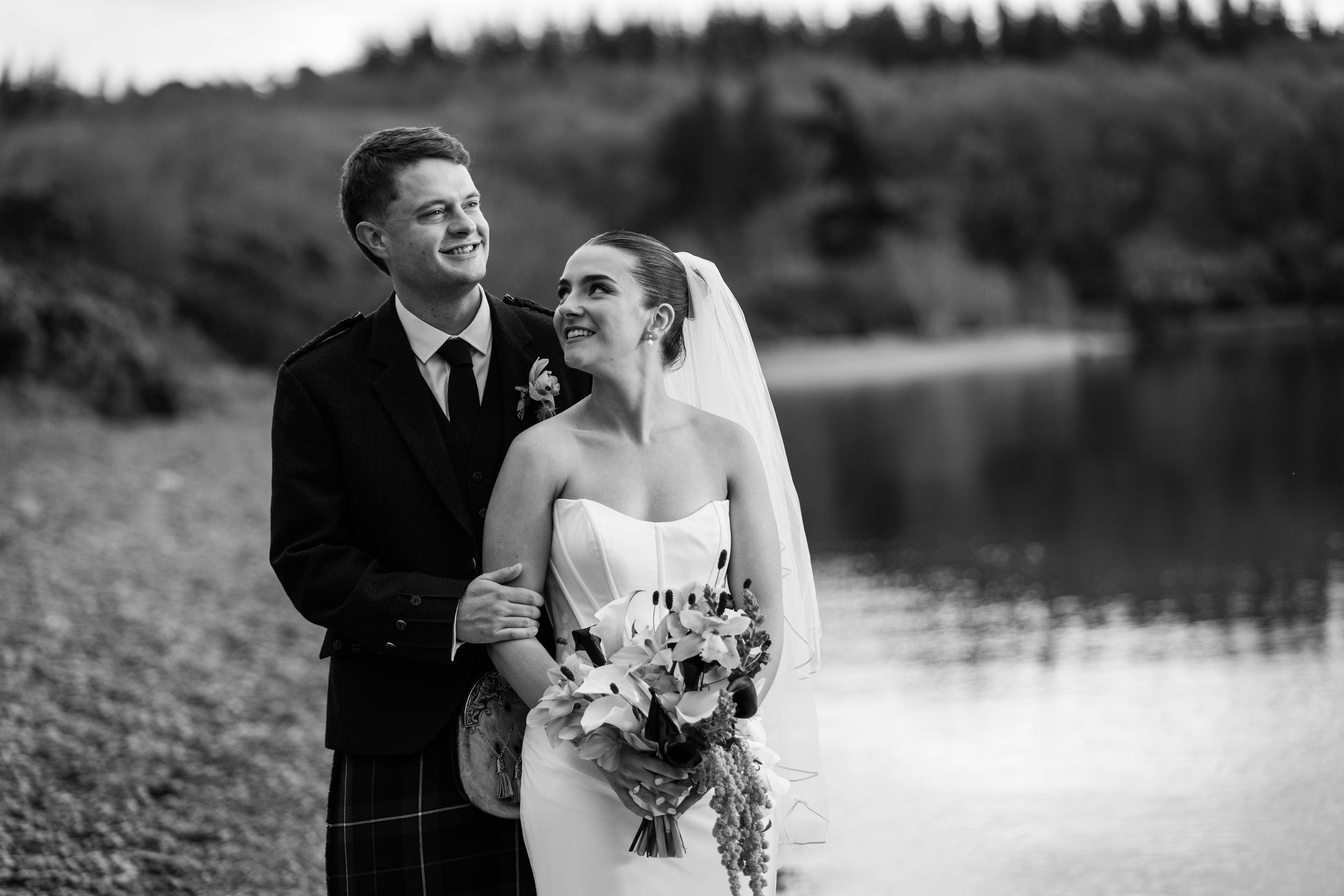 Black and white wedding photo of a smiling couple by a lake, bride holding bouquet, groom in tartan kilt.