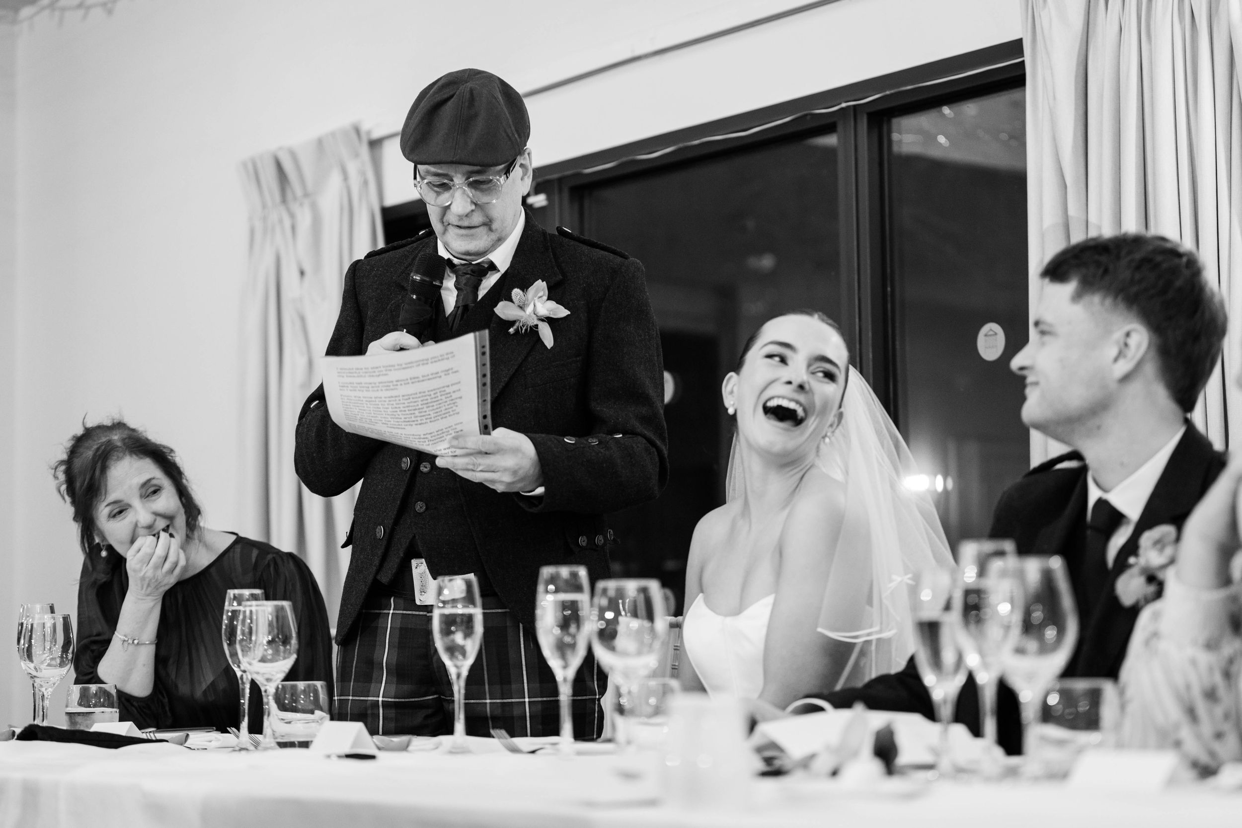 Black and white wedding reception photo of bride laughing joyfully as older man delivers humorous speech at elegant dinner.