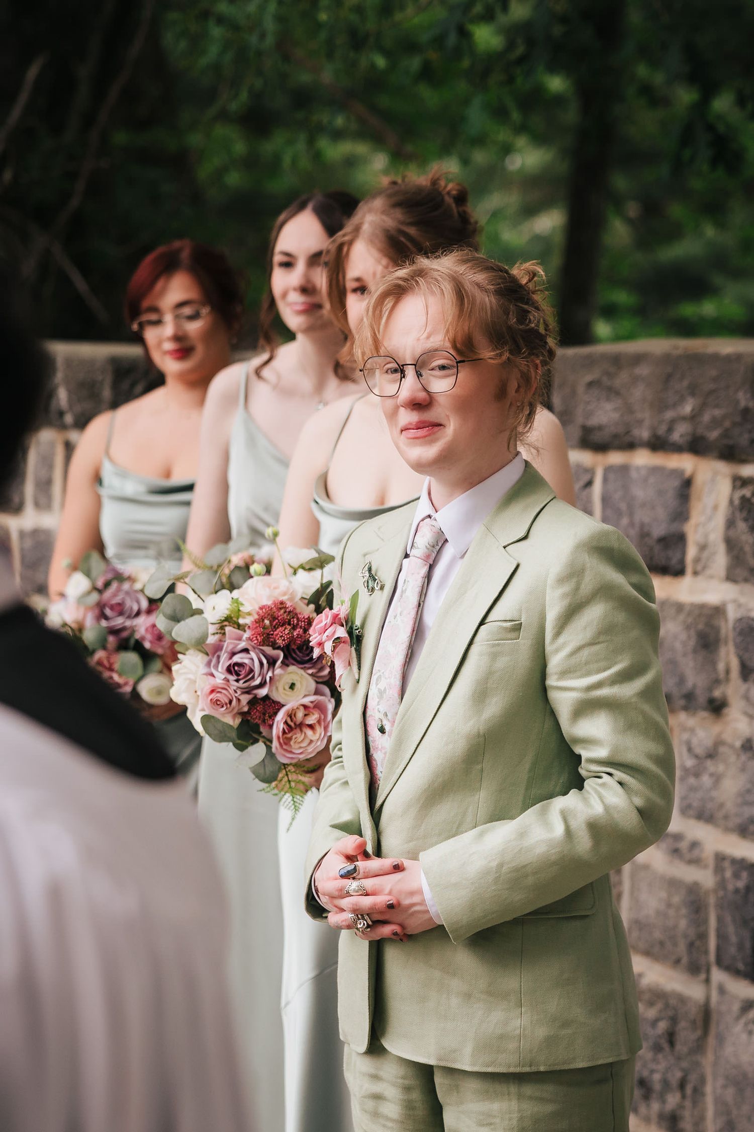 Maid of honor in a sage green suit tearing up during the ceremony, bridesmaids in matching sage dresses behind her.