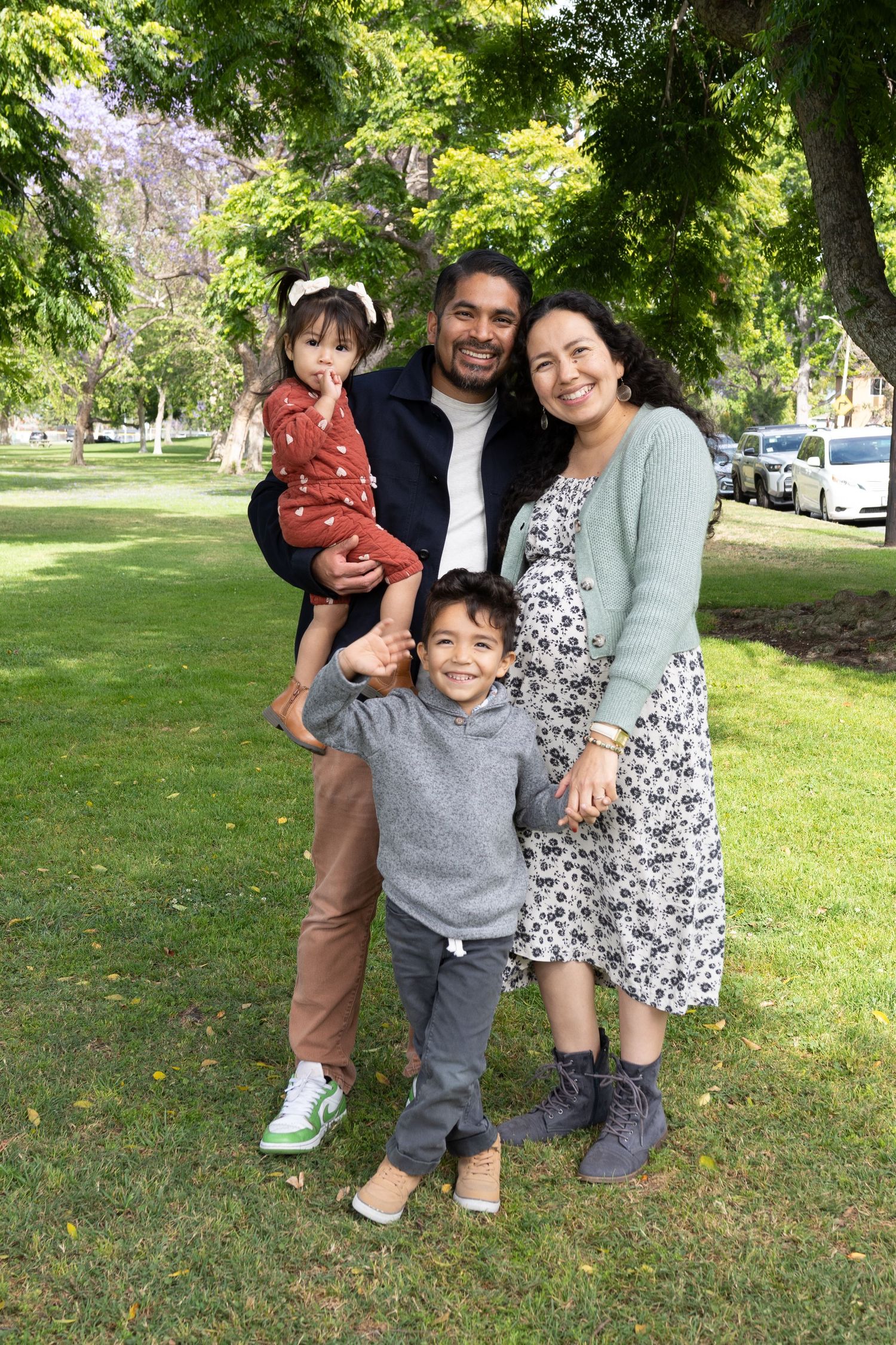 Happy family of four posing outdoors in a green park, parents smiling with two young children on a sunny day.