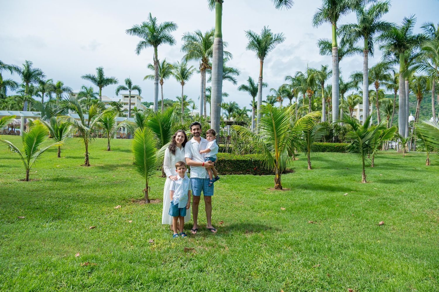 Family of four posing on lush green lawn surrounded by tall palm trees at a tropical resort.