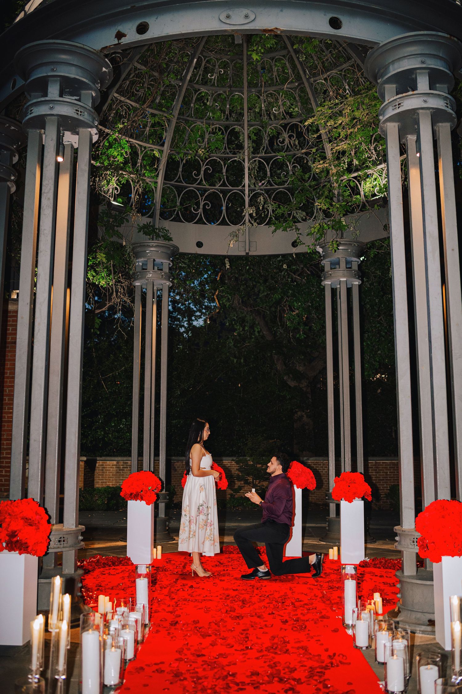 Man proposes on one knee under elegant gazebo with red rose petals, candles, and floral arrangements at night.