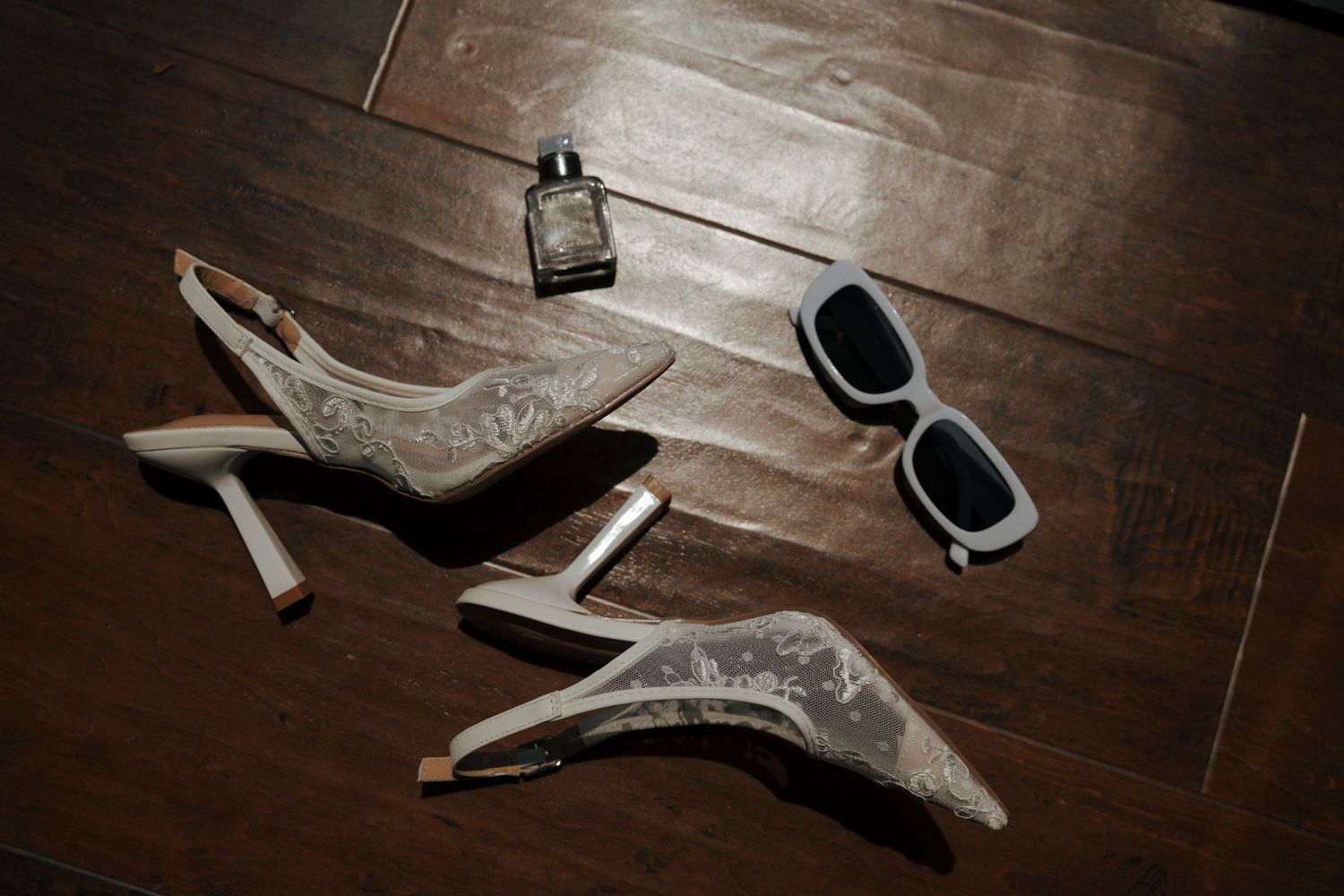 Elegant lace slingback heels, perfume bottle, and white sunglasses arranged on dark hardwood floor.