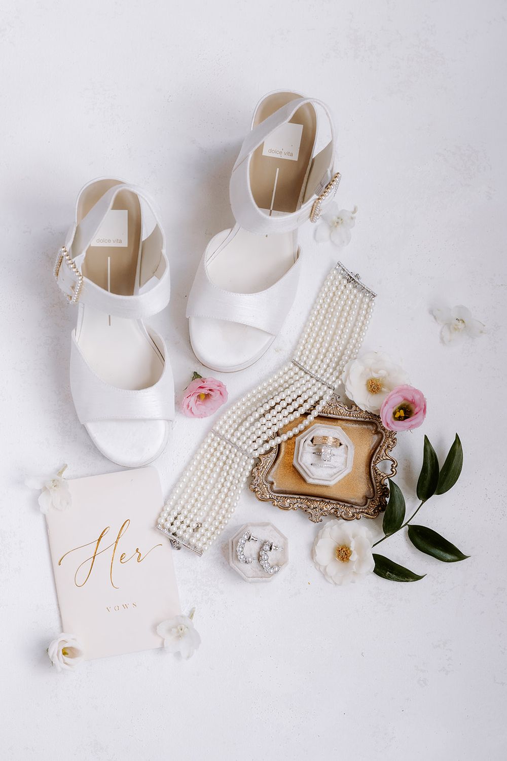White bridal heels, pearl bracelet, rings on gold tray, earrings, and pink flowers arranged on white surface.